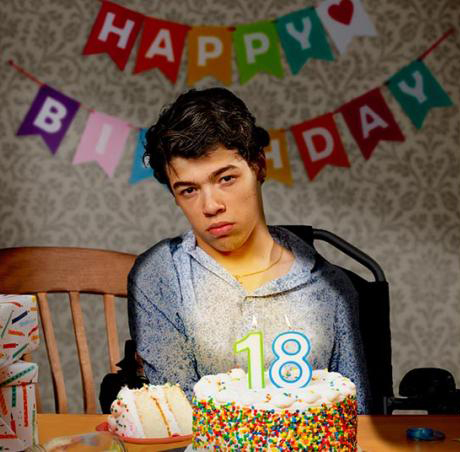 A young man in his wheelchair is sitting in front of a birthday cake with candles for his eighteenth birthday. He is wearing a blue dress shirt with a print design. He has a slice of cake on a plate in front of him and there is a happy birthday sign in the background.